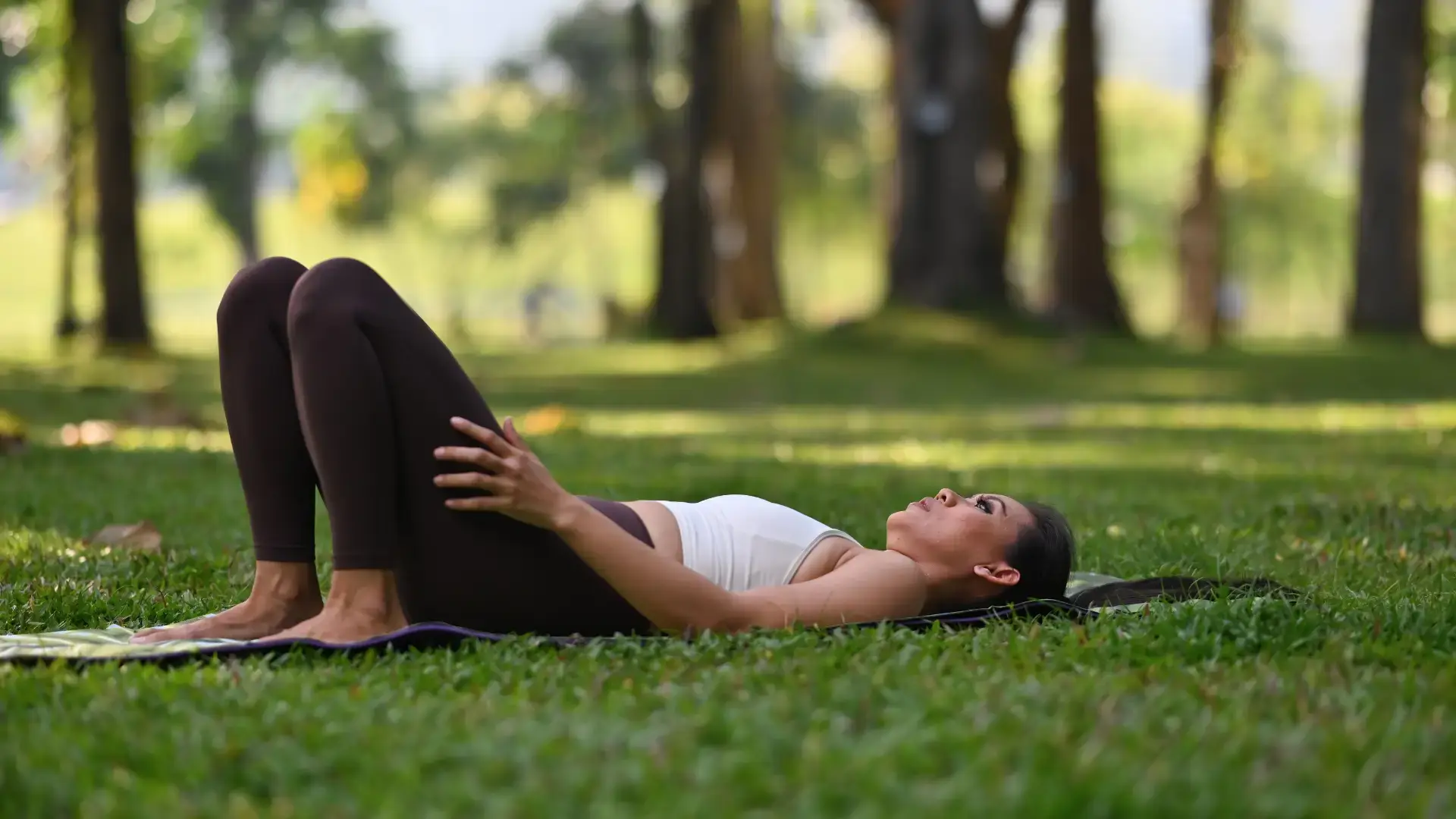 Woman lying on a mat in a park in a constructive rest position with knees bent and one hand resting on her leg, surrounded by soft green grass and trees Trauma Therapist Institute