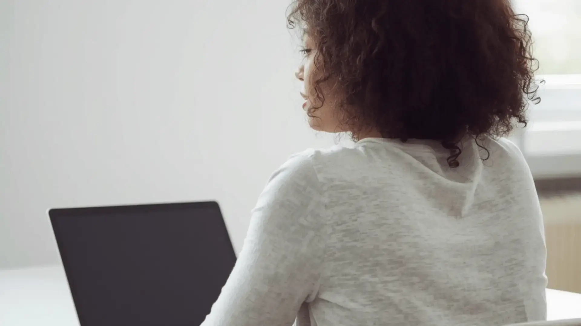 Woman seen from behind working at a laptop in a bright, minimally decorated workspace with natural light Trauma Therapist Institute