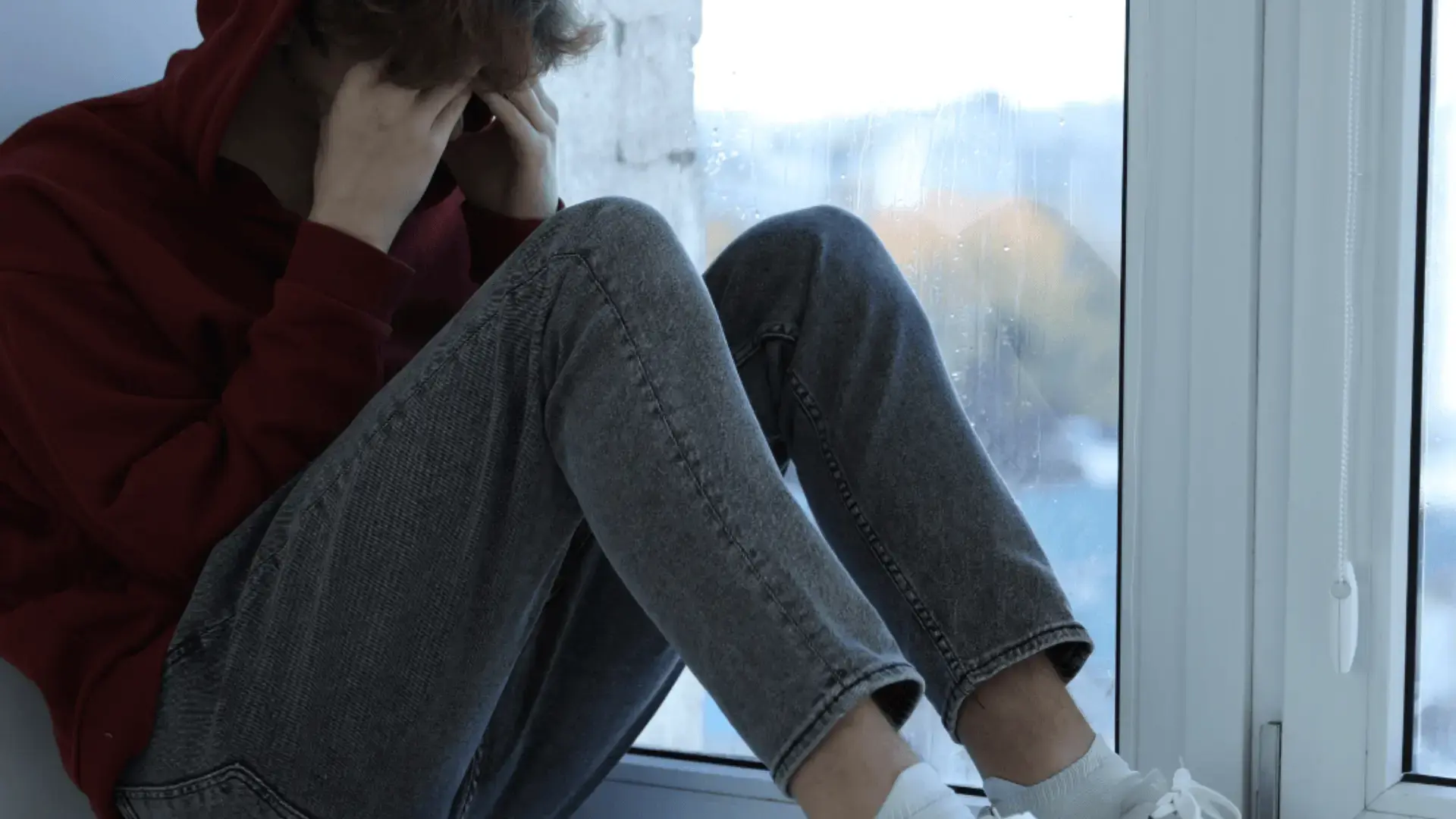 Young person in a red hoodie sitting curled up on a windowsill with their face in their hands, rain visible on the window behind them Trauma Therapist Institute