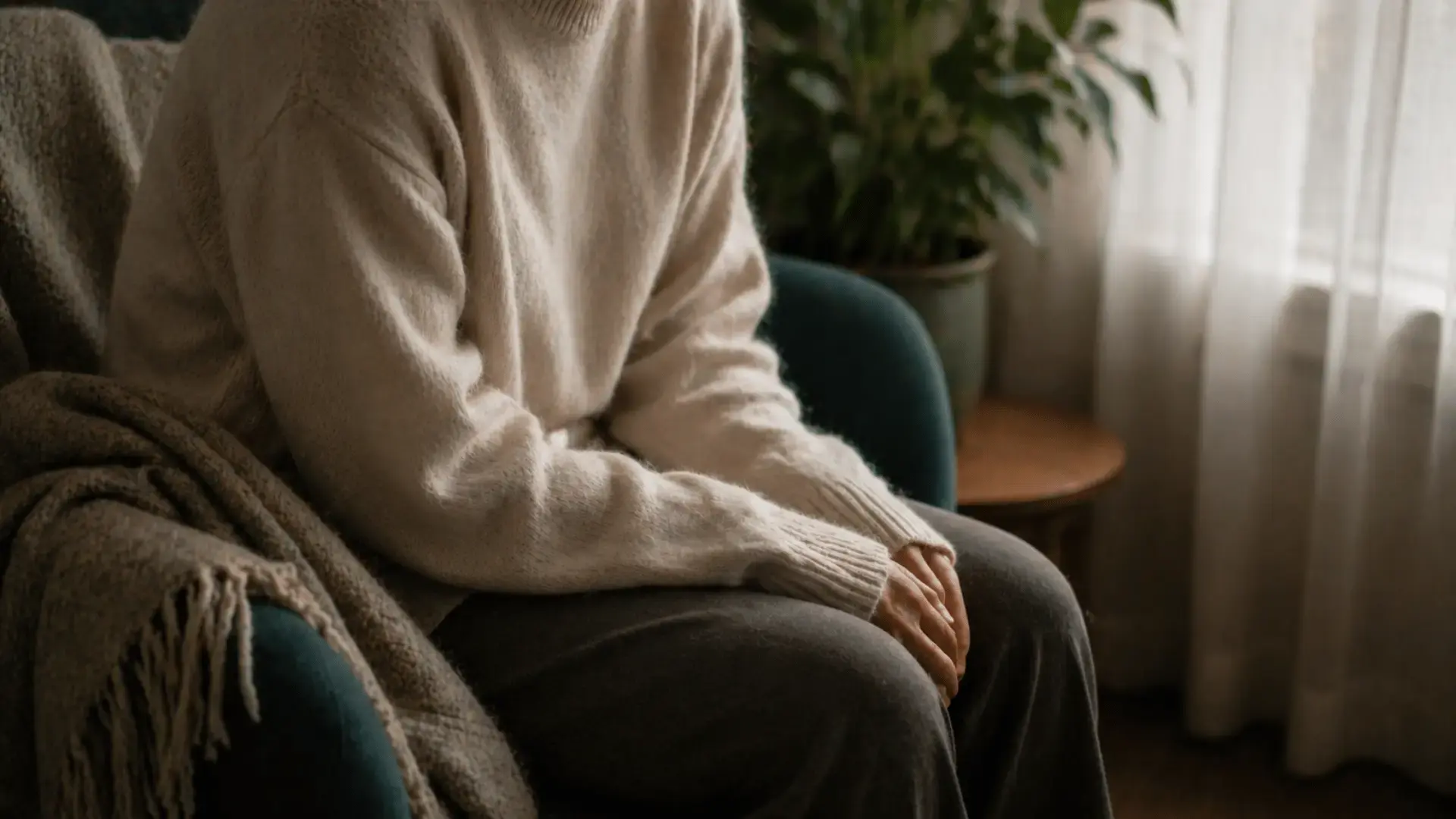 A person sits alone in a soft chair near a window with shoulders rounded forward and posture collapsed inward, body wrapped in an oversized knit sweater, illustrating the felt experience of dorsal vagal shutdown — the nervous system's freeze response of withdrawal and disconnection.